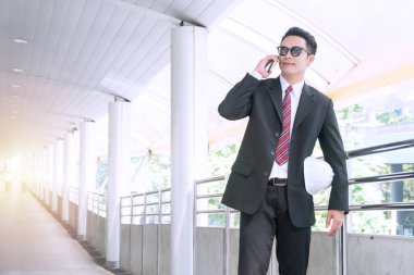Güneş gözlüğü takan yakışıklı genç işadamı elinde akıllı telefonla konuşuyor modern şehrin arka planında duran beyaz kaskı tutuyor.. 