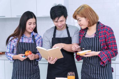 Asyalı arkadaş grupları mutfakta birlikte yemek pişiriyorlar. Neşeli gülümseyen genç adam elinde kitap tutarak evde bayan arkadaşlarıyla yemek pişirmeyi öğreniyor. Arkadaşlık kavramı.