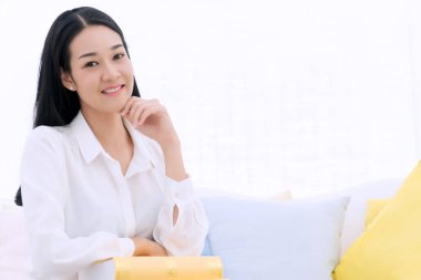 Yaşam tarzı rahatlama konsepti. Neşeli, gülümseyen genç bir kadın beyaz koltukta oturuyor. Yastık ve sarı kitapla evde oturuyor.