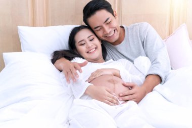 Koca, beyaz yatakta karısının karnını sevgiyle kucaklıyor. Mutlu bir çift genç adam yatak odasında hamile güzel karısına sarılıyor..