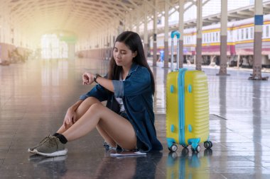 Seyahat konsepti. Sarı valizli çekici gezgin kız tren istasyonunun peronunda oturuyor. Trende bekleyen hippi genç bir kadın kamu istasyonundayken kol saatine bakıyor..