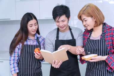 Asyalı arkadaş grupları mutfakta birlikte yemek pişiriyorlar. Neşeli gülümseyen genç adam elinde kitap tutarak evde bayan arkadaşlarıyla yemek pişirmeyi öğreniyor. Arkadaşlık kavramı.