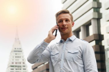 Teknoloji ve insan konsepti. Çekici bir genç adam ofis binasının dışındaki akıllı telefonuyla işten bahsediyor..