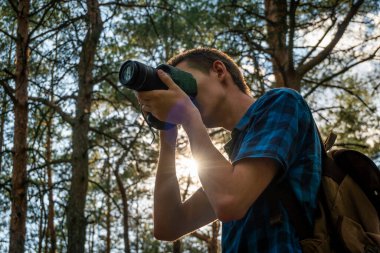 Mavi ekose gömlekli genç adam elinde kamerayla kozalaklı ormanda.