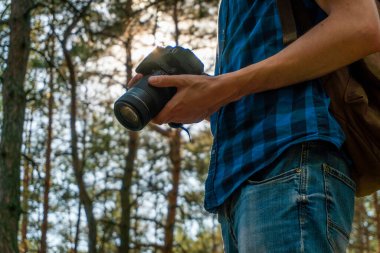 Mavi ekose gömlekli genç adam elinde kamerayla kozalaklı ormanda.