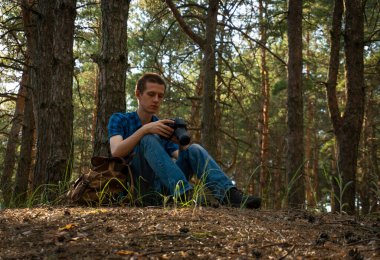 Mavi ekose gömlekli genç adam elinde kamerayla kozalaklı ormanda.