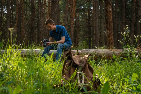 Mavi ekose gömlekli genç adam elinde kamerayla kozalaklı ormanda.