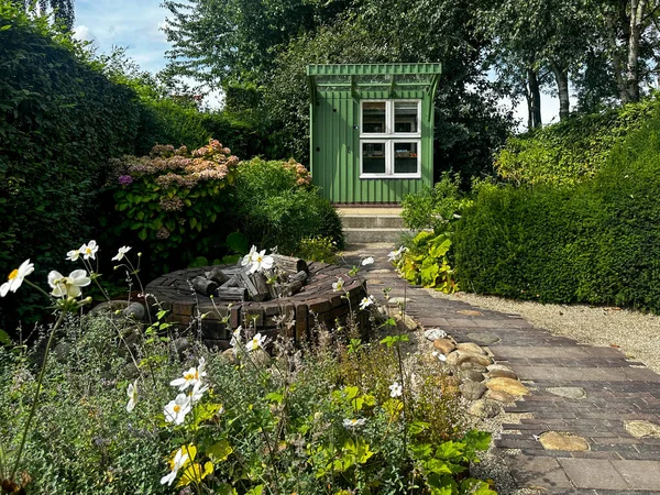 Apeltern, Hollanda, 28 Ağustos 2025: Taş yolu ve atölye olarak kullanılan yeşil kulübesi olan bir bahçe. Büyük taşlarla çevrili yuvarlak bir şömine. Beyaz şakayıklar ve ortancalar çiçek açtı