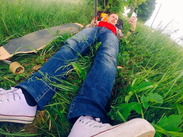 Deitado na grama, o cara está descansando depois de montar um longboard . — Fotografia de Stock