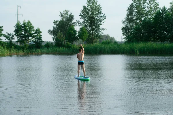 Een man staat op een bord van de Sap met een riem en zwemt door een bos meer, — Stockfoto