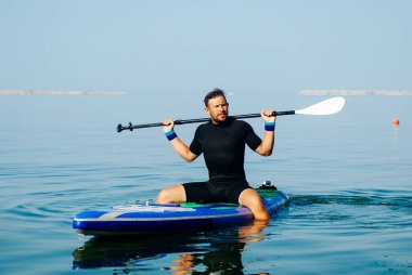 Sahilde bir Sup kurulu ile bir sörfçü portresi. Şafakta Paddleboard üzerinde oturan genç adam. Doğa sporları kavramı. Erkek sörfçü yaşam tarzı.