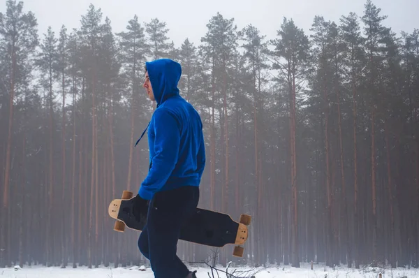 Close-up de um adolescente, de pé na camisola azul segurando a mão de um skate no fundo de uma floresta de inverno, um patinador perdido , — Fotografia de Stock
