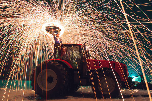Man sparks on the background of a red tractor, steel wool photo, sparks on a long exposure,