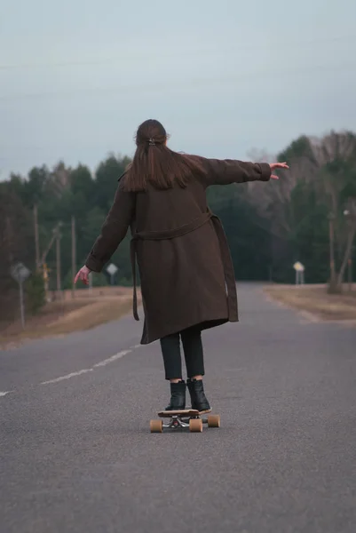Meisje model skateboarden op een landelijke weg, modieuze meisje in een lange jas op een skateboard, — Stockfoto
