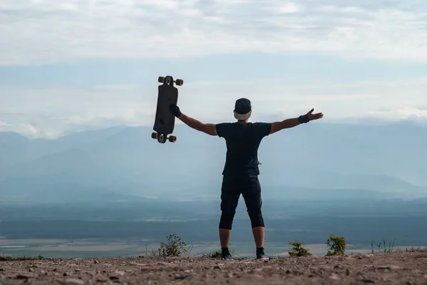 Een jonge man in een pet met een board in zijn handen staat op een klif hoog in de bergen tegen de hemel, een reis op een skateboard, — Stockfoto