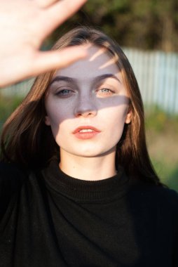 Genç bir kız eliyle yüzünü kapatıyor. Güneşin ışınları sokakta siyah giysiler giyen bir kızın yüzüne ve ellerine düşer.,