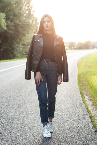 Portrait of beautiful brunette young girl in black leather jacket at sunset walking along the road, — Stock Photo, Image