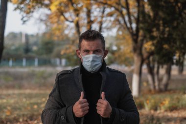 Sonbahar Parkı 'nın arka planında palto ve tıbbi maske takan bir adam. Coronavirus sonbahar dalgası konsepti.