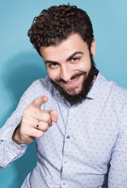 Neşeli sakallı adam onun parmak kamera işaret. Işık gömlek ve tarafsız bir mavi arka plan