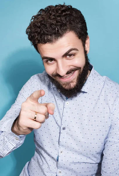 Neşeli sakallı adam onun parmak kamera işaret. Işık gömlek ve tarafsız bir mavi arka plan