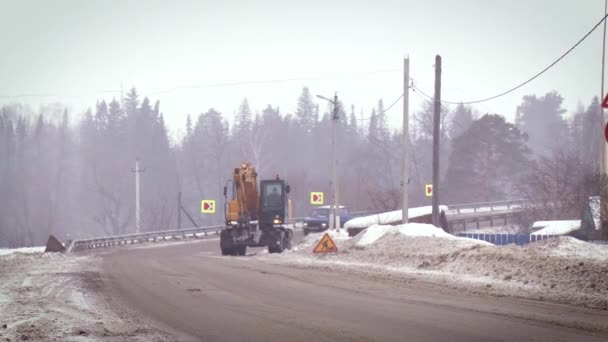 A estrada funciona. Escavadeira no processo de trabalhar em uma estrada coberta de neve contra o fundo da floresta. Dia de inverno — Vídeo de Stock