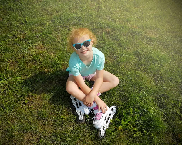 Ontspanning, kindertijd, openluchtspelen en sport concept - schattige kleine roodharige meisje zittend op groen gras in rolschaatsen en camera kijken. — Stockfoto