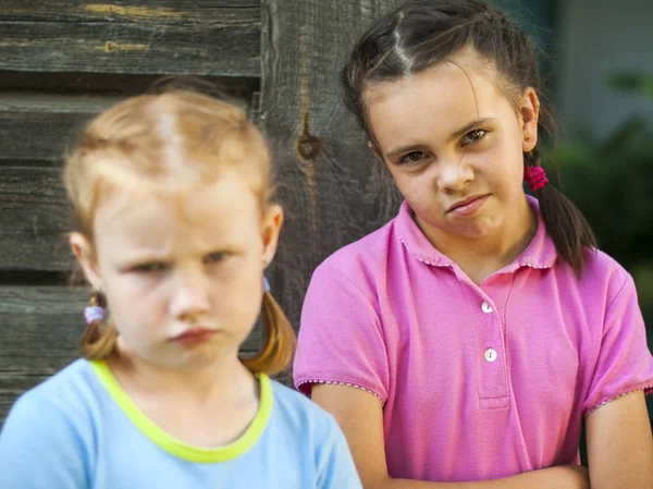 Two girls frowned, quarreled, stand on the street, focus on the far girl. Two evil and gloomy sisters are standing apart from each other, not talking to each other. Redhead and brunette. Against the background of a wooden house, summer weather. In th