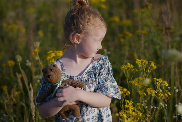 Lifestyle photo portrait of a girl outdoors in a flower field, looking into the distance. In the hands of a knitted bear, red hair gathered in a bun.