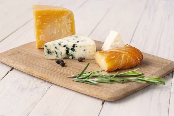 Ost av olika sorter på en trä kartong, vitt träbord. Diverse ostar på trä skärbräda. Roquefort Cheese Cheddar hård ostskivor SOF ost och rosmarin. — Stockfoto