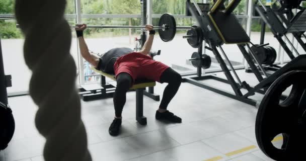 Unga idrottare gör bänkpress i gymmet. Man ligger på bänken och tränar med skivstång. Slow motion — Stockvideo