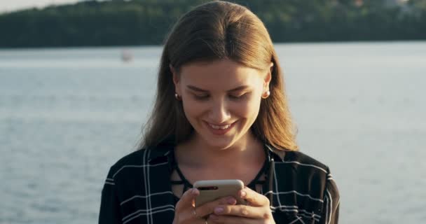 Primer plano retrato de mujer atractiva joven sonriendo y mirando el teléfono inteligente cerca de la playa al atardecer confiada belleza — Vídeo de stock