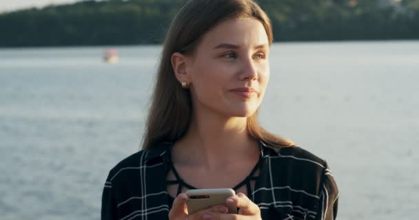 Nahaufnahme Porträt einer jungen attraktiven Frau, die bei Sonnenuntergang lächelt und ihr Smartphone in Strandnähe betrachtet — Stockvideo