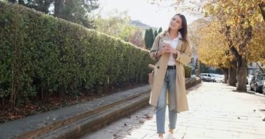 Genç çekici iş kadını, tarihi yeşil kentsel alanda geziyor, sıcak kahve içiyor, güneş ışığına bakıyor ve çalılık duvarının yanındaki iş elbisesi içinde geleceği düşünüyor.