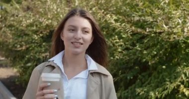 Genç çekici iş kadını, tarihi yeşil kentsel alanda geziyor, sıcak kahve içiyor, güneş ışığına bakıyor ve çalılık duvarının yanındaki iş elbisesi içinde geleceği düşünüyor.