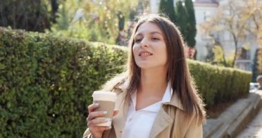 Genç çekici iş kadını, tarihi yeşil kentsel alanda geziyor, sıcak kahve içiyor, güneş ışığına bakıyor ve çalılık duvarının yanındaki iş elbisesi içinde geleceği düşünüyor.