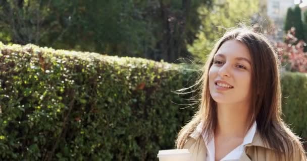 Young attractive business woman walks around historic green urban area, with take away cup and drink hot coffee, looking at sunlight and thinking about future, dressed in business suit near bush wall — Stock Video