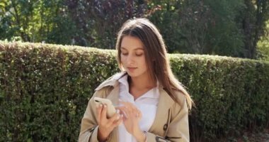 Çekici esmer kız güneşte kalıyor ve akıllı telefonunu kullanıyor, kameraya bakıyor ve gülümsüyor, güneşli bir havada.