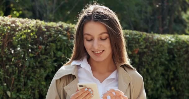 Ελκυστική μελαχρινή κοπέλα που μένει στον ήλιο και χρησιμοποιεί το smartphone της, κοιτάζοντας στην κάμερα και χαμογελώντας, ηλιόλουστο καιρό — Αρχείο Βίντεο