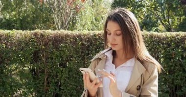 Çekici esmer kız güneşte kalıyor ve akıllı telefonunu kullanıyor, kameraya bakıyor ve gülümsüyor, güneşli bir havada.