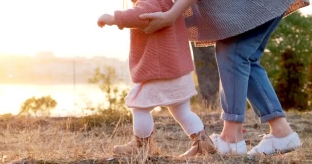 Una joven madre camina de la mano con su hija al aire libre. Familia feliz caminando sobre hierba verde al atardecer en el parque — Vídeo de stock