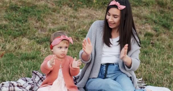 Mutter und kleine Tochter verbringen die Zeit gemeinsam in einem Stadtpark bei einem Picknick. junge Frau und kleines Mädchen spielen auf einem Plaid im grünen Gras, bei Sonnenuntergang — Stockvideo