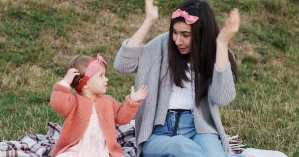 Мати і маленька дочка проводять разом час у міському парку на пікніку. Молода жінка з маленькою дівчинкою бавляться на штукатурці в зеленій траві на заході сонця. — стокове відео