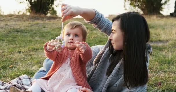 Mutter und kleine Tochter verbringen die Zeit gemeinsam in einem Stadtpark bei einem Picknick. junge Frau und kleines Mädchen spielen auf einem Plaid im grünen Gras, bei Sonnenuntergang — Stockvideo
