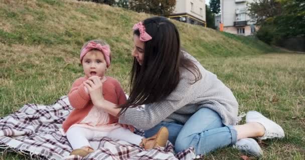 Madre y una hija pequeña, pasan tiempo juntas en un parque de la ciudad en un picnic. Mujer joven y niña están jugando en un cuadros en hierba verde, al atardecer — Vídeo de stock