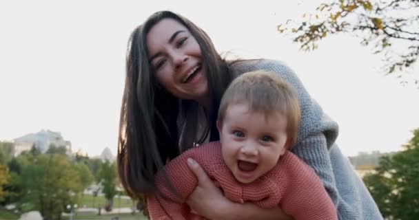 Mujer joven y bonita sosteniendo a la niña en sus brazos al atardecer en el parque. Feliz madre jugando, abrazando y besando a su pequeña hija — Vídeo de stock