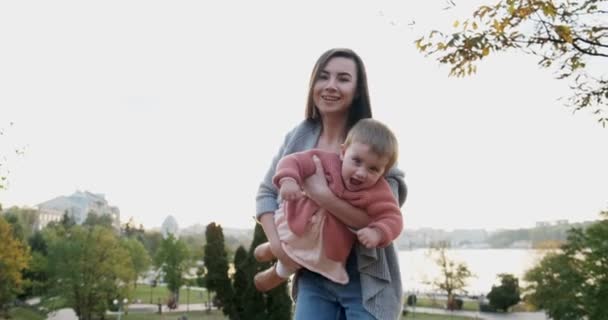 Piękna młoda kobieta trzymająca córeczkę w ramionach o zachodzie słońca w parku. Szczęśliwa matka bawiąca się, przytulająca i całująca swoją małą córkę — Wideo stockowe