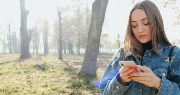 Güneşli bir sonbahar parkında gülümseyen ve akıllı telefona bakan çekici genç bir kadının yakın portresi. — Stok video