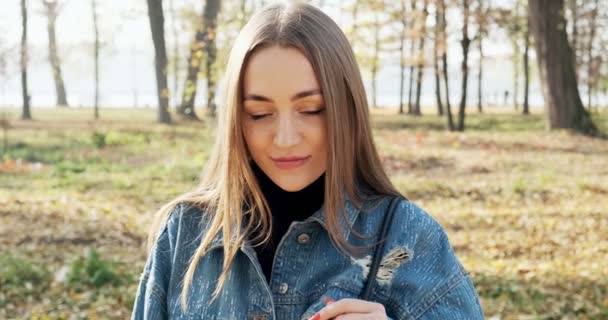 Retrato de una joven atractiva hembra que sonríe y mira a la cámara en el parque de otoño. Alegre mujer bonita en el clima soleado. De cerca. — Vídeo de stock