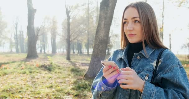 Крупный план портрета молодой привлекательной женщины, улыбающейся и смотрящей на смартфон в солнечном осеннем парке — стоковое видео