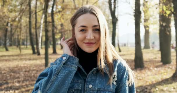 Retrato de una joven atractiva hembra que sonríe y mira a la cámara en el parque de otoño. Alegre mujer bonita en el clima soleado. De cerca. — Vídeos de Stock